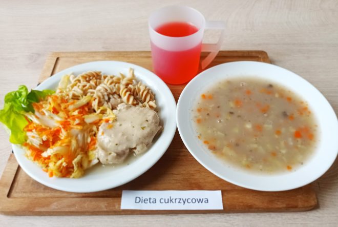 Na drewnianej tacy ustawiono pełnowartościowy obiad dla osoby na diecie cukrzycowej. Po lewej stronie znajduje się talerz z dwoma rodzajami pełnoziarnistego makaronu świderki, surówką z białej kapusty i marchwi na liściu sałaty oraz kawałkiem duszonego mięsa w jasnym sosie ziołowym. Obok talerza stoi przezroczysty kubek z czerwonym kompotem, prawdopodobnie słodzonym w sposób dostosowany do diety. Po prawej stronie – talerz z porcją zupy jarzynowej z widocznymi kawałkami ziemniaków, marchwi, selera i kaszy. Posiłek jest starannie przygotowany i estetycznie podany, a przed tacą widnieje etykieta z napisem „Dieta cukrzycowa”
