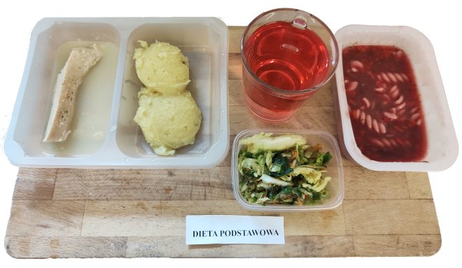 Zestaw obiadowy na drewnianej tacy, oznaczony jako „Dieta podstawowa”, podany w pojemnikach jednorazowych. Po lewej stronie znajduje się dwudzielne naczynie – w jednej przegródce kawałek gotowanego mięsa (najpewniej filet z kurczaka) w jasnym, rzadkim sosie, w drugiej – dwie porcje ziemniaków puree o puszystej konsystencji. Obok, w przezroczystym kubku, znajduje się napój w intensywnie czerwonym kolorze – prawdopodobnie kompot owocowy. Po prawej stronie leży podłużny pojemnik z zupą buraczkową z drobnym makaronem typu świderki. Na przodzie tacy stoi niewielki plastikowy pojemnik z surówką – mieszanka kapusty pekińskiej, marchwi i zielonych warzyw liściastych. Całość skomponowana jest z myślą o zbilansowanym posiłku dla pacjentów bez specjalnych ograniczeń żywieniowych, przy zachowaniu zasad prostoty i strawności