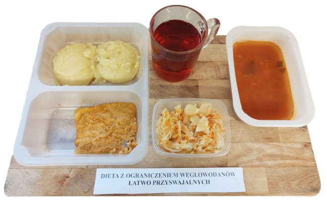 Zestaw obiadowy w diecie z ograniczeniem węglowodanów łatwo przyswajalnych, podany na tacy z podziałką. W górnym lewym przegródce znajdują się dwie kulki purée ziemniaczanego. W dolnej części dania głównego leży panierowany filet rybny o złocistym kolorze. Obok w przezroczystym pojemniku znajduje się porcja surówki z kiszonej kapusty z dodatkiem marchwi i cebuli. Po prawej stronie widoczna miseczka z czerwono-pomarańczową zupą jarzynową z drobnymi kawałkami warzyw oraz szklanka z herbatą owocową o rubinowej barwie. Całość umieszczona na drewnianej tacy z etykietą informującą o rodzaju diety: „DIETA Z OGRANICZENIEM WĘGLOWODANÓW ŁATWO PRZYSWAJALNYCH”