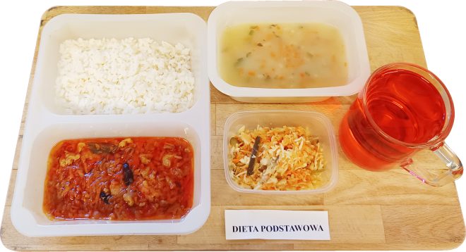 Na drewnianej desce ustawiono zestaw obiadowy w ramach "Diety podstawowej". W plastikowym pojemniku dwukomorowym znajduje się porcja białego ryżu oraz sos z warzywami i mięsem w czerwonym sosie pomidorowym. Obok, w osobnym pojemniku, znajduje się jasna zupa jarzynowa z widocznymi kawałkami marchewki i zieleniny. Na desce ustawiono także małe pudełko z surówką z kapusty i marchewki oraz szklankę z czerwonym napojem owocowym. Przy zestawie leży karteczka z napisem "Dieta podstawowa"