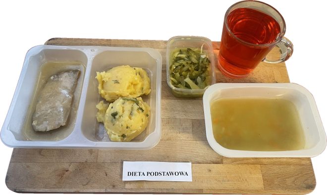 Na drewnianej tacy znajduje się obiad zgodny z założeniami diety podstawowej. W plastikowym pojemniku z trzema przegródkami podano gotowany kawałek mięsa w jasnym sosie, porcję puree ziemniaczanego z koperkiem oraz duszoną fasolkę szparagową z cebulą w osobnym, przezroczystym pudełku. Po prawej stronie – głęboki pojemnik z przezroczystą zupą jarzynową o delikatnym kolorze, z widocznymi fragmentami warzyw. Do zestawu dołączono szklankę z czerwonym napojem, najprawdopodobniej herbatą owocową lub kompotem. Całość podana estetycznie, z etykietą „DIETA PODSTAWOWA” umieszczoną przed tacą