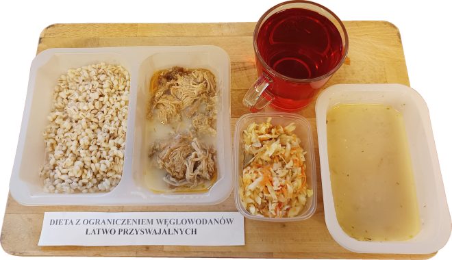Na drewnianej desce ułożono zestaw obiadowy w plastikowych pojemnikach. W lewym pojemniku znajduje się porcja gotowanego pęczaku, w środkowym – szarpane mięso drobiowe w lekkim sosie. Obok, w oddzielnym pojemniku, surówka z białej kapusty z dodatkiem marchewki. Po prawej stronie – jasna, klarowna zupa o konsystencji bulionu. Z tyłu szklanka z czerwonym naparem owocowym. Na karteczce wydrukowany napis: „Dieta z ograniczeniem węglowodanów łatwo przyswajalnych”