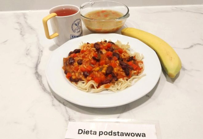 Na białym talerzu znajduje się porcja makaronu z warzywnym sosem z czerwoną fasolą, kukurydzą, marchewką i papryką. Obok szklana miska z klarowną zupą warzywną. Zestaw uzupełnia dojrzały banan oraz kubek z herbatą owocową, podany w białym kubku z żółtym uchem, ozdobionym logo sanatorium. Całość oznaczona jako „Dieta podstawowa”. Posiłek został podany na marmurowym blacie, utrzymanym w czystej i jasnej estetyce