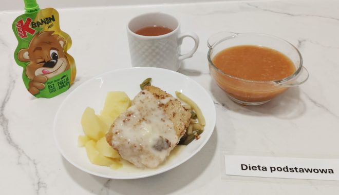 Kompletny zestaw obiadowy na białym blacie. Na głównym talerzu znajduje się panierowany kotlet schabowy z jasnym sosem, ziemniaki gotowane i mieszanka warzyw z fasolki szparagowej. Z boku – przezroczysta miska z zupą pomidorową lub jarzynową na czerwonym wywarze. Obok biały kubek z herbatą oraz saszetka musu jabłkowo-bananowego w tubce z rysunkiem misia. Na stole leży etykieta „Dieta podstawowa”