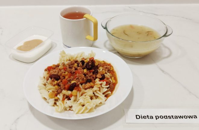 Zestaw obiadowy ustawiony na białym blacie. W centrum talerz z makaronem świderki oraz gulaszem z mielonego mięsa, czerwonej fasoli, kukurydzy i marchewki w sosie pomidorowym. Po prawej stronie szklana miska z zupą ziemniaczaną lub jarzynową na bazie wywaru, z widocznymi nitkami koperku. W tyle kubek z herbatą owocową i żółtym uchem. Po lewej stronie plastikowy pojemnik z deserem – jogurt naturalny z dodatkiem musu jabłkowego lub karmelowego. Na dole karteczka z napisem „Dieta podstawowa”