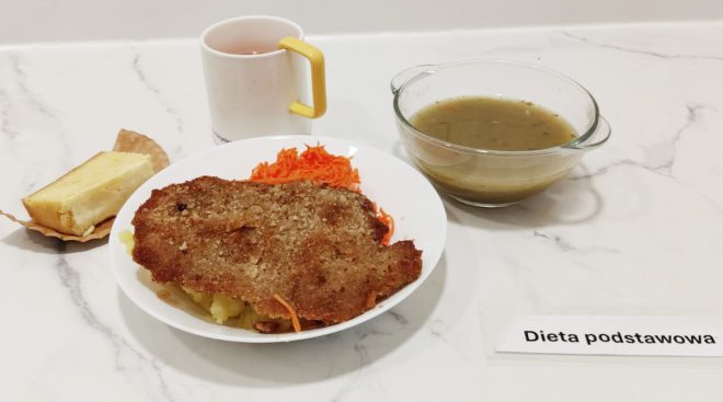 Kotlety schabowe w chrupiącej panierce na gotowanych ziemniakach, obok surówka z tartej marchewki. Na talerzu także kawałek ciasta serowego. W tle kubek z napojem oraz miseczka z zupą – prawdopodobnie jarzynową lub ziemniaczaną. Na stole widoczna kartka z napisem „Dieta podstawowa”