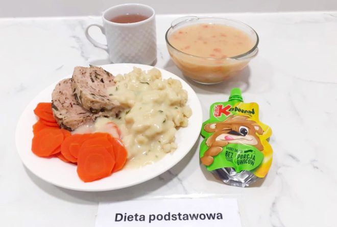 Pełnowartościowy obiad w diecie podstawowej serwowany na białym talerzu: dwa plastry pieczeni wieprzowej w ziołach, polane delikatnym sosem, podane z kluskami kładzionymi i gotowaną marchewką w plasterkach. W tle znajduje się miseczka z zupą jarzynową w kremowej, pomarańczowej zalewie, kubek z herbatą oraz mus owocowy w saszetce o smaku jabłkowo-bananowym z wizerunkiem misia. Wszystko ustawione na jasnym marmurowym blacie z etykietą "Dieta podstawowa"
