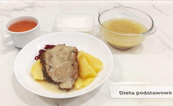 Na talerzu obiadowym podano plaster pieczeni wieprzowej w sosie, ułożony na gotowanych ziemniakach. Obok znajduje się porcja czerwonej kapusty zasmażanej. Do zestawu dołączono przezroczystą miskę z jasnym bulionem oraz niewielki pojemnik z białym sosem, prawdopodobnie śmietanowym. Na stole znajduje się także filiżanka z herbatą. Wszystkie elementy ułożone są na jasnym, marmurowym blacie. W prawym dolnym rogu widnieje etykieta „Dieta podstawowa”