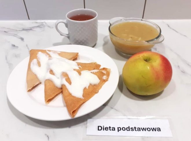 Posiłek w ramach „Diety podstawowej” złożony z trzech naleśników złożonych w trójkąty, polanych białym jogurtem lub śmietaną, ułożonych na talerzu. Obok – czerwono-zielone jabłko, kubek z ciemną herbatą oraz miseczka z gęstym kompotem owocowym lub zupą owocową. Wszystko ustawione na marmurowym blacie