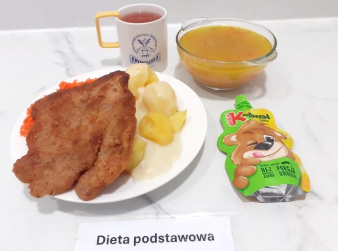 Na zdjęciu widnieje zestaw obiadowy w ramach diety podstawowej, starannie ułożony na białym, marmurkowym blacie. Na głównym talerzu znajduje się duży panierowany kotlet schabowy o apetycznie zrumienionej skórce, porcja gotowanych ziemniaków oblanych delikatnym jasnym sosem oraz surówka z tartej marchewki. Obok ustawiono przezroczystą szklaną miseczkę z gorącą zupą jarzynową o złocistej barwie i widocznymi kawałkami warzyw. Z prawej strony leży owocowy mus Kubuś 4xOwoc o smaku jabłkowym, przeznaczony jako deser – opakowanie zdobi wizerunek uśmiechniętego misia. Do posiłku dołączono kubek z herbatą owocową, z charakterystycznym logotypem uzdrowiska „Kryniczanka”. Całość opatrzona jest etykietą z napisem „Dieta podstawowa”