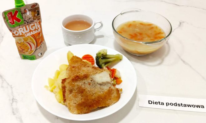 Na marmurowym blacie ustawiono obiad w ramach "Diety podstawowej". Na talerzu znajdują się gotowane ziemniaki, gotowane warzywa (brokuł, marchew, kalafior) oraz panierowany filet rybny. Obok talerza stoi przezroczysta miska z zupą jarzynową z kawałkami marchwi. Zestaw uzupełnia biały kubek z różowym naparem oraz saszetka musu owocowego "Kubuś Drugie Śniadanie" o smaku jabłko-mango. Przy potrawach widoczna kartka z napisem "Dieta podstawowa"