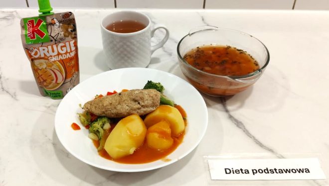 Na drewnianej desce ustawiono zestaw obiadowy w ramach "Diety podstawowej". W plastikowym pojemniku dwukomorowym znajduje się porcja białego ryżu oraz sos z warzywami i mięsem w czerwonym sosie pomidorowym. Obok, w osobnym pojemniku, znajduje się jasna zupa jarzynowa z widocznymi kawałkami marchewki i zieleniny. Na desce ustawiono także małe pudełko z surówką z kapusty i marchewki oraz szklankę z czerwonym napojem owocowym. Przy zestawie leży karteczka z napisem "Dieta podstawowa"