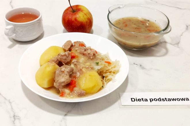 Na marmurowym blacie znajduje się zestaw obiadowy oznaczony etykietą „Dieta podstawowa”. W centrum ustawiono głęboki, biały talerz z porcją gotowanych ziemniaków, gulaszem wieprzowym z marchewką w jasnym sosie oraz gotowaną kiszoną kapustą z natką pietruszki. W tle znajduje się szklana miseczka z zupą jarzynową – lekkim wywarem z widocznymi kawałkami warzyw. Obok niej – czerwone jabłko oraz filiżanka z napojem przypominającym herbatę. Kompozycja posiłku jest zgodna z wytycznymi żywienia szpitalnego, zbilansowana i estetycznie podana