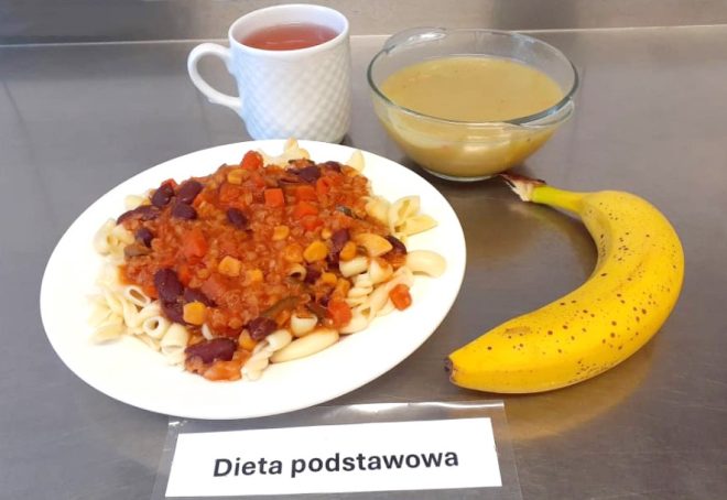 Na stalowym blacie ułożono zestaw obiadowy. Na talerzu znajduje się makaron rurki z warzywnym sosem pomidorowym z fasolą i kukurydzą. Obok miska z zupą kremową oraz kubek z herbatą. Całość dopełnia banan. Przed talerzem umieszczono kartkę z napisem „Dieta podstawowa”
