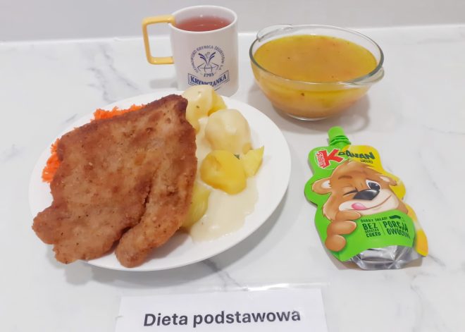 Zdjęcie przedstawia pełnowartościowy obiad serwowany w ramach diety podstawowej. Na centralnym, białym talerzu ułożony jest duży panierowany kotlet schabowy, podsmażony na złoto. Obok znajduje się porcja gotowanych ziemniaków z lekkim sosem oraz surówka z tartej marchewki. W tle widoczna jest szklana miseczka z jasną, klarowną zupą – prawdopodobnie jarzynową. Z prawej strony talerza znajduje się owocowy mus Kubuś o smaku jabłkowym w saszetce. Do zestawu dołączono kubek z herbatą owocową – biały z żółtym uchem, z logiem uzdrowiska "Kryniczanka". Całość sfotografowano na jasnym, marmurkowym blacie. Przed potrawami widnieje etykieta z napisem „Dieta podstawowa”
