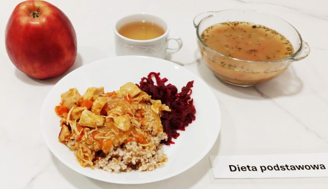 Na marmurowym blacie ustawiono zestaw obiadowy z oznaczeniem „Dieta podstawowa”. W centrum kompozycji znajduje się biały talerz z porcją kaszy gryczanej, na której ułożono gulasz z kawałkami duszonego kurczaka, plasterkami marchewki i cebuli w lekkim, pomarańczowym sosie. Obok leży porcja duszonych buraczków o intensywnie bordowym kolorze. Po lewej stronie talerza stoi duże, czerwone jabłko o błyszczącej skórce. W tle po prawej widoczna jest przezroczysta miseczka z klarowną zupą – najpewniej jarzynową, z kawałkami warzyw i ziołami pływającymi na powierzchni. Towarzyszy jej filiżanka z herbatą, ustawiona centralnie w tle. Całość wygląda estetycznie i apetycznie, prezentując zbilansowany i lekkostrawny posiłek obiadowy, przygotowany z myślą o podstawowej diecie szpitalnej lub sanatoryjnej