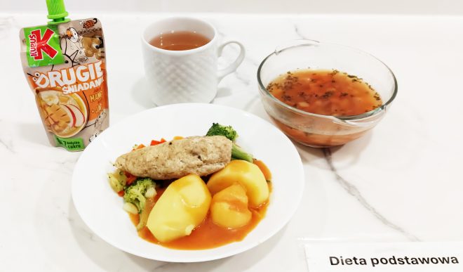 Na białym blacie znajduje się zestaw obiadowy oznaczony jako "Dieta podstawowa". Na pierwszym planie widać biały talerz z porcją gotowanych ziemniaków polanych czerwonym sosem, porcją gotowanych warzyw (brokuły, marchew, kalafior) oraz kotletem drobiowym o jasnobrązowej barwie. Obok talerza znajduje się przezroczysta miska wypełniona klarowną zupą jarzynową z widocznymi kawałkami warzyw i ziołami. W tle ustawiono biały kubek z herbatą oraz saszetkę musu owocowego "Kubuś Drugie Śniadanie" o smaku jabłko-mango. Całość ułożona jest estetycznie na jasnym marmurowym blacie, pod którym widoczna jest karteczka z napisem "Dieta podstawowa"
