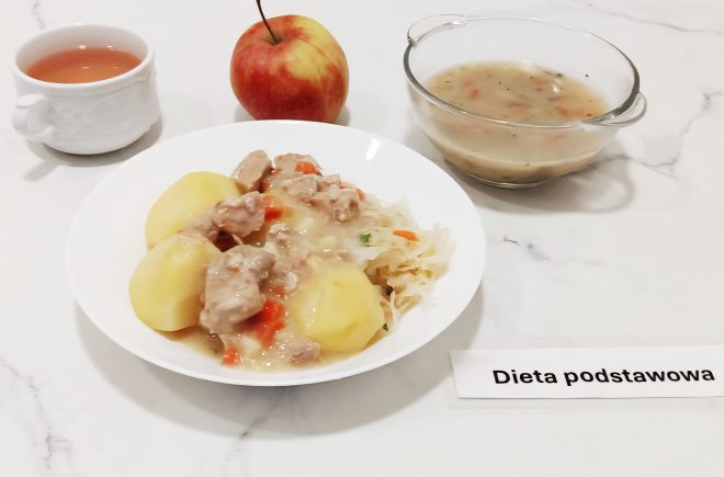 a marmurowym blacie ustawiono zestaw obiadowy oznaczony jako „Dieta podstawowa”. Na środku znajduje się biały talerz z ziemniakami gotowanymi w całości, kawałkami duszonego mięsa w jasnym sosie z kawałkami marchewki oraz porcją gotowanej kiszonej kapusty. Obok, w szklanej miseczce, podano porcję zupy jarzynowej na bazie bulionu z dodatkiem ziół. W tle widoczne są: czerwone jabłko oraz filiżanka z jasnym napojem – prawdopodobnie herbatą. Posiłek wygląda na zbilansowany i przygotowany z myślą o codziennym żywieniu szpitalnym