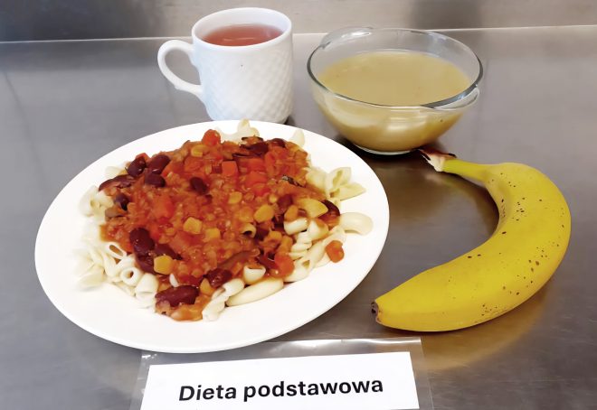 Zestaw obiadowy ułożony na stalowym blacie. Na talerzu makaron rurki z sosem warzywnym na bazie pomidorów, fasoli i kukurydzy. W tle kubek z herbatą oraz miska z zupą o kremowej, jasnożółtej konsystencji. Z boku dołączony banan. Na kartce nadrukowany napis „Dieta podstawowa”