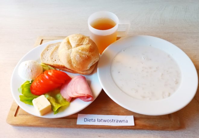Na jasnym, drewnianym blacie przygotowano śniadanie w ramach "Diety łatwostrawnej". Na białym talerzu ułożono bułkę typu kajzerka, dwa cienkie kromki chleba pszennego, kilka plastrów pomidora, liść sałaty, plasterek szynki, porcję masła oraz kulkę białego twarogu. Obok talerza znajduje się biały głęboki talerz wypełniony jasną, kremową zupą mleczną z drobną kaszą lub płatkami. W tle widoczny jest półprzezroczysty kubek z jasnym napojem owocowym. Całość ustawiona na drewnianej tacy z karteczką "Dieta łatwostrawna"
