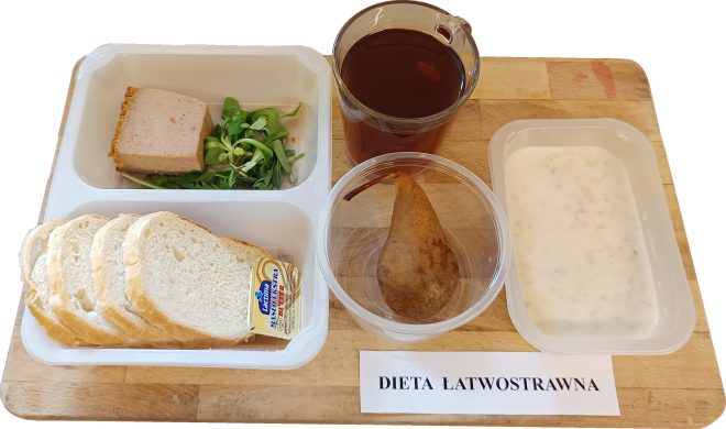 Śniadanie w diecie łatwostrawnej: jasne pieczywo (cztery kromki), kostka masła oraz porcja pasztetu z zieleniną (rukolą lub roszponką) na białej tacko-talerzyku. Obok: połówka gruszki w przezroczystym pojemniku, czarny napój (kawa zbożowa lub herbata) w szklance oraz miseczka z jasną zupą mleczną z dodatkiem płatków. Wszystko ułożone na desce z napisem „DIETA ŁATWOSTRAWNA”