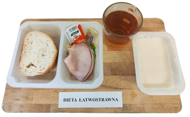 Śniadanie w wersji łatwostrawnej, podane na drewnianej tacy w pojemnikach jednorazowych, oznaczone etykietą „Dieta łatwostrawna”. W jednej przegródce znajduje się kromka jasnego pieczywa pszennego. Obok – w drugiej części pojemnika – porcja dodatków: plasterki wędliny drobiowej na liściu sałaty, porcja masła, mały kubeczek dżemu truskawkowego oraz listek z dodatkiem warzywnym (najprawdopodobniej ogórek lub sałata). Po prawej stronie ustawiony jest kubek z ciepłą herbatą, a w oddzielnym naczyniu – lekka kaszka mleczna o kremowej konsystencji. Kompozycja śniadania odpowiada potrzebom osób na diecie lekkostrawnej – bez ostrych przypraw i ciężkostrawnych składników, z naciskiem na łagodne, neutralne smaki i wartości odżywcze