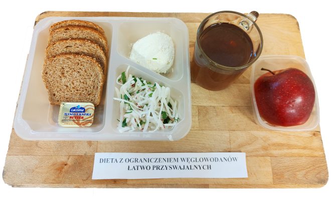 Śniadanie w wersji diety z ograniczeniem łatwo przyswajalnych węglowodanów, starannie ułożone na plastikowej tacy. Po lewej stronie znajdują się cztery kromki pieczywa pełnoziarnistego oraz porcja masła „Lacima Masło Ekstra”. W środkowej przegródce leży kula twarogu z dodatkiem szczypiorku, poniżej – surówka ze świeżych warzyw: rzodkwi, natki pietruszki i odrobiny marchwi. Po prawej stronie na tacy szklanka z herbatą ziołową oraz oddzielnie zapakowane jabłko odmiany Red Delicious o błyszczącej, czerwonej skórce. Na drewnianej podstawie umieszczona etykieta z nazwą diety: „DIETA Z OGRANICZENIEM WĘGLOWODANÓW ŁATWO PRZYSWAJALNYCH”