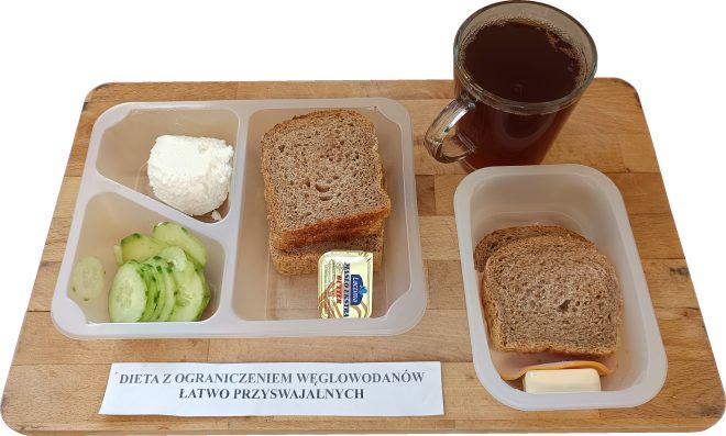 Na drewnianej tacy zaprezentowano śniadanie przygotowane zgodnie z założeniami diety z ograniczeniem węglowodanów łatwo przyswajalnych. W plastikowym pojemniku znajdują się trzy przegródki – w jednej ułożono kulkę chudego twarogu, w drugiej plasterki świeżego ogórka, a w trzeciej kilka kromek razowego chleba oraz porcję masła w plastikowym opakowaniu. Obok, w oddzielnym pojemniku, znajduje się kanapka z pełnoziarnistego pieczywa z dodatkiem chudej wędliny i sera żółtego. Do zestawu dołączono szklankę z ciemnym napojem, najpewniej kawą zbożową lub herbatą bez cukru. Przed tacą widnieje etykieta z napisem „DIETA Z OGRANICZENIEM WĘGLOWODANÓW ŁATWO PRZYSWAJALNYCH”