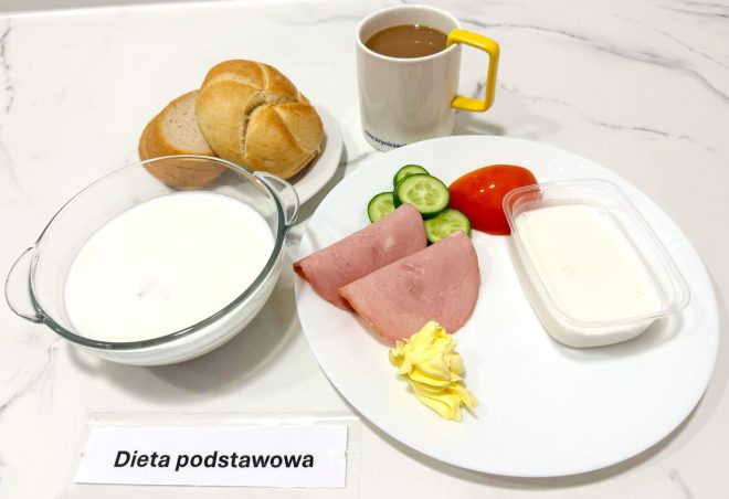 Na talerzu znajdują się dwa plastry szynki drobiowej, kilka plasterków świeżego ogórka i pomidora, porcja masła oraz deser mleczny w plastikowym pojemniczku. Obok widzimy miseczkę z mlekiem i pieczywo (bułka pszenna i kromka chleba razowego). Do posiłku podano również kawę z mlekiem. Wszystko serwowane na jasnym blacie, oznaczone jako „Dieta podstawowa”