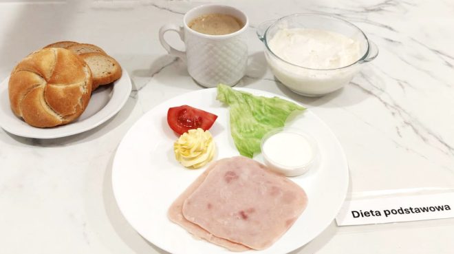 Śniadanie zgodne z dietą podstawową, elegancko podane na marmurowym blacie. Na dużym białym talerzu znajdują się dwa cienkie plastry szynki drobiowej, liść sałaty lodowej, plasterek pomidora, niewielka porcja masła uformowana w ozdobną rozetę oraz mały kubeczek z jogurtem naturalnym. Obok – miseczka z białym twarożkiem o gładkiej konsystencji. Na osobnym talerzyku po lewej stronie – bułka kajzerka oraz kromka chleba mieszanego. Całość uzupełnia biała filiżanka z kawą z mlekiem lub zbożową. Zestaw oznaczony kartką z napisem „Dieta podstawowa”