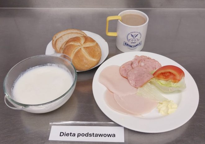 Tradycyjne śniadanie w diecie podstawowej. Na talerzu kilka plasterków wędliny (w tym drobiowej i wieprzowej), kawałek sałaty lodowej, ćwiartka pomidora oraz porcja masła. W tle filiżanka z kawą zbożową, bułka i pieczywo pszenne oraz miska z białym napojem mlecznym (np. kefirem lub mlekiem). Wszystko podane na metalicznym blacie z etykietą „Dieta podstawowa”
