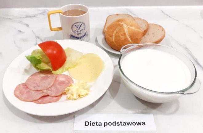 Śniadanie w diecie podstawowej składające się z kilku plasterków szynki wieprzowej, dwóch plasterków sera żółtego, masła oraz plasterka pomidora i liścia sałaty. Obok znajduje się filiżanka z kawą z mlekiem, miska z białym mlekiem lub kefirem, a także bułka kajzerka i kromki chleba pszennego. Wszystko podane na marmurowym blacie z karteczką informacyjną "Dieta podstawowa"