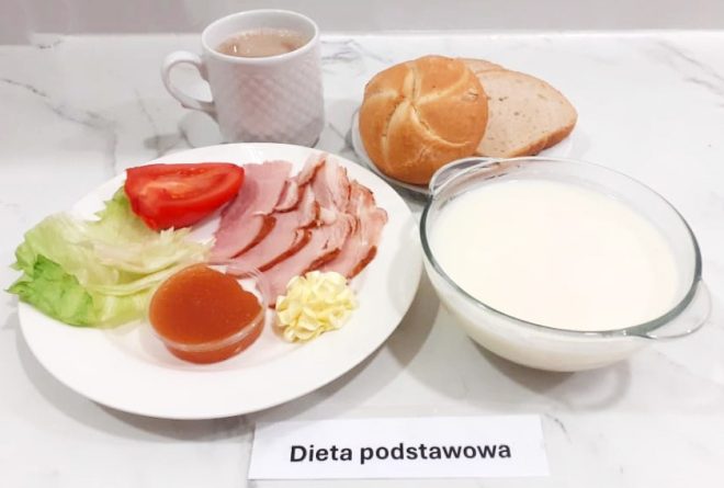 Zestaw śniadaniowy opisany jako „Dieta podstawowa” podany na białym marmurowym blacie. Na talerzu znajdują się plasterki wędliny, sałata lodowa, cząstka pomidora, porcja masła i galaretki owocowej. Obok talerza stoi filiżanka z kawą z mlekiem, a z tyłu położono kajzerkę i kromkę pieczywa razowego. Całość dopełnia miseczka z białym jogurtem naturalnym lub mlekiem