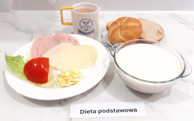 Na śniadaniowym zestawie diety podstawowej widzimy bogaty i zbilansowany posiłek. Na białym talerzu ułożono dwa plasterki wędliny drobiowej, dwa plastry żółtego sera, świeżą ćwiartkę pomidora, listek sałaty oraz rozetkę z masła. Obok, w osobnym naczyniu, znajduje się duża miska z białym napojem mlecznym lub kefirem. W tle widoczny jest talerzyk z pieczywem – jedną bułką kajzerką oraz dwoma kromkami jasnego chleba pszennego. Kubek z napojem (najpewniej kawa zbożowa z mlekiem) opatrzony jest logiem „Kryniczanka” i żółtym uchem. Wszystko ustawione zostało na jasnym blacie, a pod kompozycją umieszczono kartkę z napisem „Dieta podstawowa”