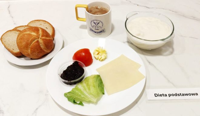 Na marmurowym blacie przygotowano śniadanie oznaczone jako "Dieta podstawowa". Na talerzu ułożono plasterek pomidora, liść sałaty lodowej, porcję masła w formie rozety, dwa plastry sera żółtego oraz mały kubeczek z ciemną konfiturą. Obok na talerzyku leży kilka kromek białego pieczywa oraz bułka typu kajzerka. W tle kubek z napojem (prawdopodobnie kawa z mlekiem) i szklana miska z porcją białego serka lub jogurtu. Całość uzupełnia karteczka "Dieta podstawowa"