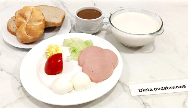 Zestaw śniadaniowy na marmurowym blacie, oznaczony jako „Dieta podstawowa”. W centrum znajduje się talerz z dwoma jajkami ugotowanymi na twardo, trzema plasterkami mielonki drobiowej, kawałkiem sałaty lodowej, plastrem czerwonego pomidora oraz dekoracyjnie podaną porcją masła. W tle – talerz z pieczywem: jedna bułka typu kajzerka i kilka kromek jasnego chleba pszennego. Obok talerzy znajduje się filiżanka z gorącym napojem, prawdopodobnie kawą zbożową lub herbatą, a po prawej – przezroczysta miseczka wypełniona mlekiem lub jogurtem naturalnym. Całość wygląda na klasyczne, lekkostrawne śniadanie w ramach diety podstawowej