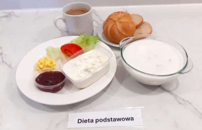 Zestaw śniadaniowy na marmurowym blacie. Na talerzu widać porcję twarogu, liść sałaty, plaster pomidora, porcję masła i dżem w małym pojemniku. W tle bułka i dwie kromki jasnego chleba. Obok szklana miseczka z jogurtem lub mlekiem z dodatkami oraz kubek z kawą z mlekiem. Na karteczce napis: „Dieta podstawowa”