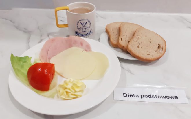 Na zdjęciu znajduje się śniadanie przygotowane w ramach diety podstawowej. Na pierwszym planie widzimy biały talerz z kilkoma plasterkami wędliny drobiowej i żółtego sera, ułożonymi obok siebie. Obok nich leży listek sałaty, ćwiartka pomidora oraz niewielka porcja masła w dekoracyjnej formie. W tle, na osobnym talerzyku, znajdują się trzy kromki jasnego chleba. Uzupełnieniem zestawu jest biały kubek z żółtym uchem i logotypem „Kryniczanka” – prawdopodobnie z kawą zbożową lub mlekiem. Kompozycję umieszczono na jasnym marmurkowym blacie. U dołu zdjęcia widnieje etykieta z napisem „Dieta podstawowa”