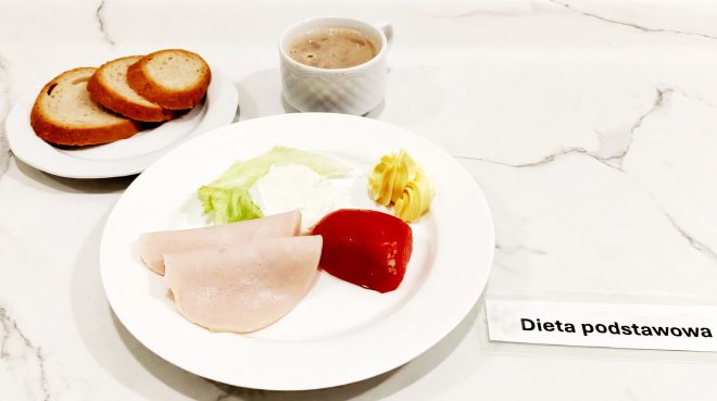 Zestaw śniadaniowy na jasnym marmurowym blacie oznaczony etykietą „Dieta podstawowa”. W centrum widnieje biały talerz z dwiema cienkimi plastrami wędliny z indyka lub szynki drobiowej, jasnym listkiem sałaty, plastrem czerwonej papryki, porcją białego twarożku i dekoracyjnym rulonikiem masła. Tło dopełnia filiżanka z kawą zbożową z mlekiem lub innym ciepłym napojem mlecznym. Obok, po lewej stronie, na osobnym spodeczku ułożono trzy kromki chleba pszennego lub mieszanego – lekko przypieczone i miękkie w środku. Całość podana w sposób schludny i zgodny z wymogami diety podstawowej, przeznaczonej dla osób bez specjalnych ograniczeń żywieniowych, lecz wymagających lekkostrawnych, klasycznych dań o wysokiej wartości odżywczej