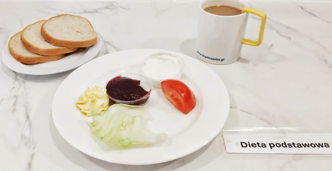 Na jasnym marmurowym blacie przygotowano śniadanie w ramach "Diety podstawowej". Na białym talerzu ułożono kawałek liścia sałaty lodowej, plaster pomidora, porcję białego serka w plastikowym kubeczku, porcję czerwonej konfitury oraz porcję masła w kształcie dekoracyjnej rozety. Obok talerza znajduje się mały talerzyk z trzema kromkami białego chleba. W tle widoczny jest biały kubek z żółtym uchem wypełniony kawą z mlekiem, na którym nadrukowano stronę internetową "kryniczanka.pl". Na blacie leży także karteczka z napisem "Dieta podstawowa"