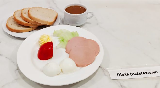 Na marmurowym blacie znajduje się zestaw śniadaniowy oznaczony etykietą „Dieta podstawowa”. Na pierwszym talerzu, na pierwszym planie, ułożono dwa jajka na twardo, trzy plasterki mielonki drobiowej, kawałek sałaty lodowej, plaster pomidora oraz porcję masła w dekoracyjnej formie rozety. W tle widoczny jest oddzielny talerz z trzema kromkami jasnego pieczywa pszennego oraz bułką kajzerką. Całości dopełnia filiżanka z gorącym napojem – prawdopodobnie kawa zbożowa lub herbata. Posiłek prezentuje się schludnie i odpowiada typowym założeniom zbilansowanej diety szpitalnej