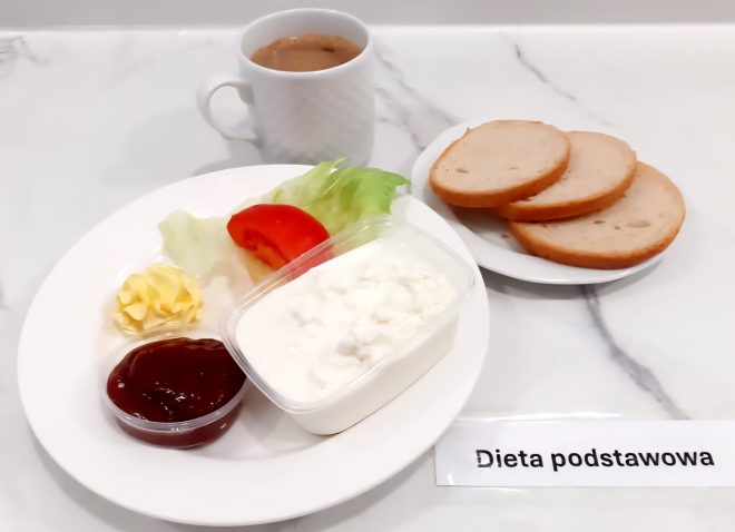 Na marmurowym blacie znajduje się talerz śniadaniowy z białym serem w plastikowym pojemniczku, listkiem sałaty i plastrem pomidora, porcją masła oraz małym pojemnikiem z dżemem. W tle kubek z kawą z mlekiem. Obok, na osobnym spodku, trzy kromki jasnego pieczywa. Na stole karteczka z napisem „Dieta podstawowa”
