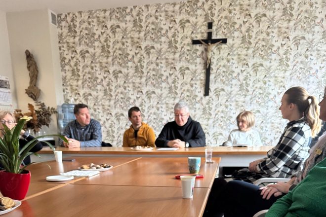 Wnętrze jasnej sali spotkań, w której przy dużym, drewnianym stole w kształcie litery „U” siedzi kilkanaście osób uczestniczących w rozmowie. W centrum kadru, przy ścianie ozdobionej tapetą z motywami roślinnymi i ptakami, siedzi starszy zakonnik w czarnym habicie – trzyma ręce splecione na blacie i mówi, skupiając uwagę pozostałych uczestników. Po jego lewej stronie siedzą dwaj mężczyźni – jeden w żółtej bluzie, drugi w koszuli w kratę, obaj uważnie słuchają. Po prawej stronie zakonnika – starsza kobieta w jasnym swetrze oraz kilka innych uczestniczek, w tym młoda kobieta w koszuli w czarno-białą kratę, siedząca bokiem do aparatu. Na pierwszym planie, na stole, widoczne są talerzyki z ciasteczkami, kubki z napojami oraz doniczka z zieloną rośliną w czerwonej osłonce. W tle, nad mówiącym zakonnikiem, wisi duży krzyż. Po lewej stronie ściany widoczna jest figura świętej oraz kalendarz i dystrybutor z wodą. Zdjęcie oddaje atmosferę skupienia i dialogu w kameralnym, przyjaznym otoczeniu