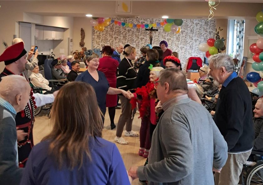 Sala wypełniona seniorami i opiekunami bawiącymi się w rytm muzyki – uczestnicy wydarzenia, ubrani w kolorowe stroje, trzymają się za ręce i tańczą w kręgu. Pomieszczenie udekorowane balonami i serpentynami, w tle kolorowy łańcuch i tapeta w liście oraz ptaki. Widoczne radosne twarze i ciepła atmosfera wspólnej zabawy