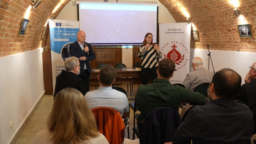 Sala konferencyjna o sklepionym ceglanym suficie. Przy stole prezydialnym siedzi zakonnik w czarnym habicie, przemawiający do mikrofonu. Obok stoi kobieta w okularach i biało-czarnej bluzie w paski, również trzymająca mikrofon. Za nimi rozwieszone są dwa roll-upy: jeden z logo projektu TECHHEALTH i drugi z logo Bonifraterskiego Centrum Medycznego