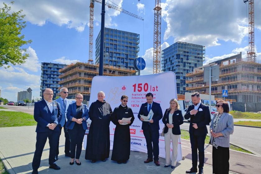 Grupa przedstawicieli Zakonu Bonifratrów, władz miasta i partnerów instytucjonalnych podczas uroczystości z okazji jubileuszu 150-lecia Szpitala i Konwentu Zakonu Bonifratrów pw. św. Aniołów Stróżów w Katowicach. W tle nowoczesna zabudowa miejska oraz rozwijające się inwestycje budowlane. Uczestnicy stoją na tle białego baneru z czerwonym napisem „150 lat”, symbolicznie podkreślającego historyczne dziedzictwo i współczesny rozwój. Atmosfera wydarzenia jest oficjalna i uroczysta, z dominacją ciemnych garniturów, habitów i słonecznego, wiosennego światła