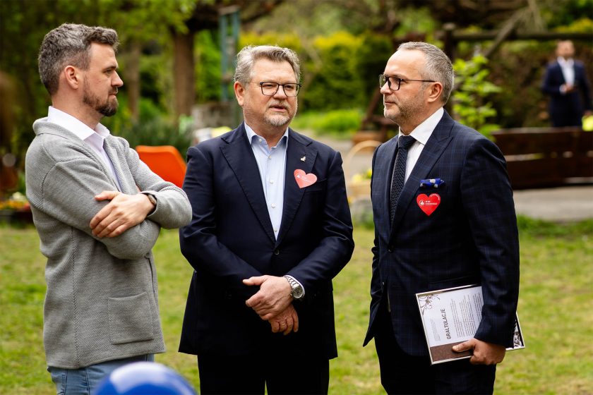 Trzej mężczyźni stoją w ogrodzie podczas wydarzenia. Dwóch z nich w garniturach z czerwonymi sercami przypiętymi do klap, jeden w swetrze z założonymi rękami. Jeden z mężczyzn trzyma w ręku ozdobny dyplom z napisem „GRATULACJE”. Tło stanowi zieleń i rozmyte sylwetki gości