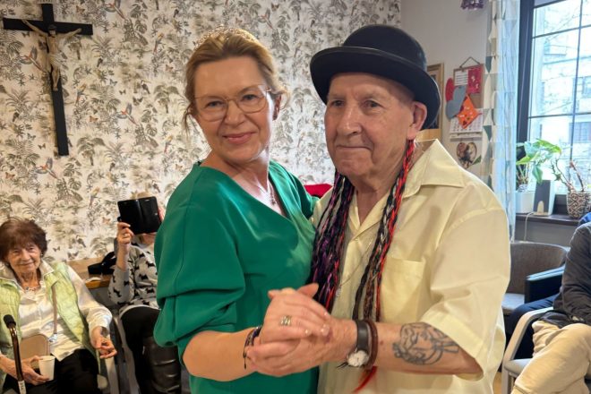 W pierwszym planie para seniorów tańczy wspólnie – kobieta w zielonej sukience z tiarą na głowie i mężczyzna w meloniku z kolorowymi warkoczykami, ubrany w jasną koszulę, z tatuażem na przedramieniu. Uśmiechnięci i skupieni na tańcu. W tle inne seniorki siedzą na krzesłach, jedna z nich fotografuje telefonem. Na ścianie za nimi widoczny jest duży krzyż z ukrzyżowanym Chrystusem oraz tapeta w motywy roślinne i ptasie