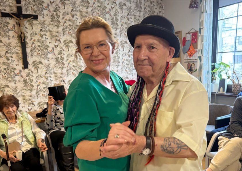 W pierwszym planie para seniorów tańczy wspólnie – kobieta w zielonej sukience z tiarą na głowie i mężczyzna w meloniku z kolorowymi warkoczykami, ubrany w jasną koszulę, z tatuażem na przedramieniu. Uśmiechnięci i skupieni na tańcu. W tle inne seniorki siedzą na krzesłach, jedna z nich fotografuje telefonem. Na ścianie za nimi widoczny jest duży krzyż z ukrzyżowanym Chrystusem oraz tapeta w motywy roślinne i ptasie