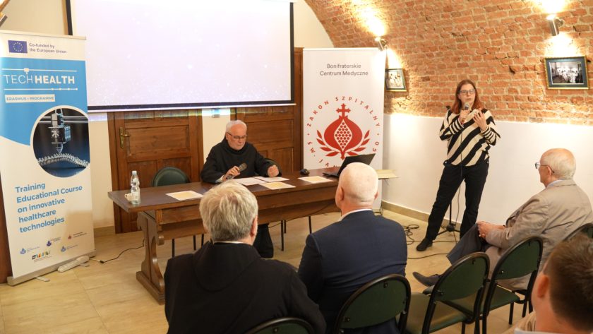 Zakonnik siedzący za stołem prezydialnym przemawia do mikrofonu. Przed nim rozłożone są dokumenty i butelka wody. Po obu stronach stołu stoją roll-upy promujące projekt TECHHEALTH i Bonifraterskie Centrum Medyczne